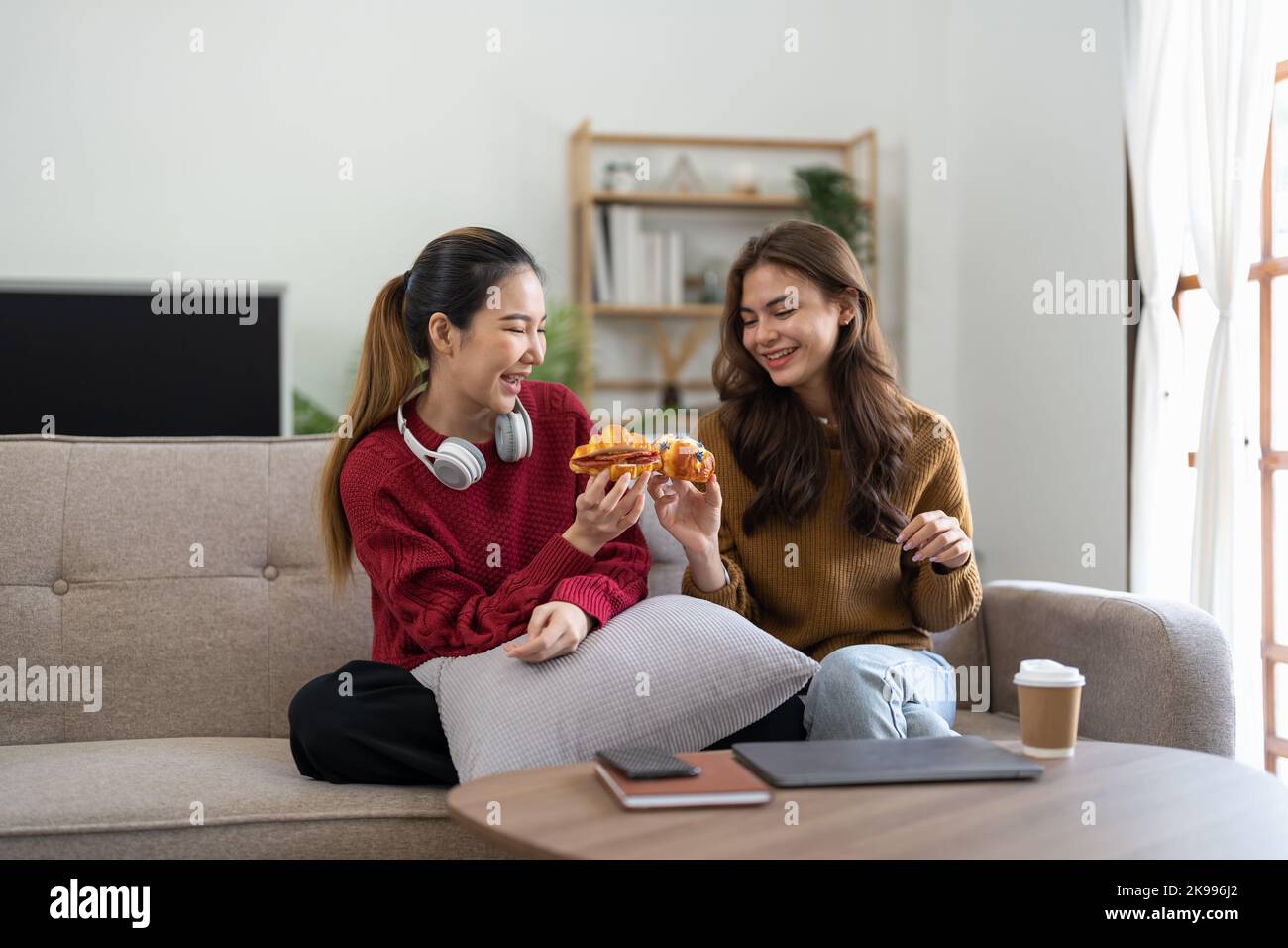 Two friends enjoying eating croissant sitting on a couch in the living room at home Stock Photo