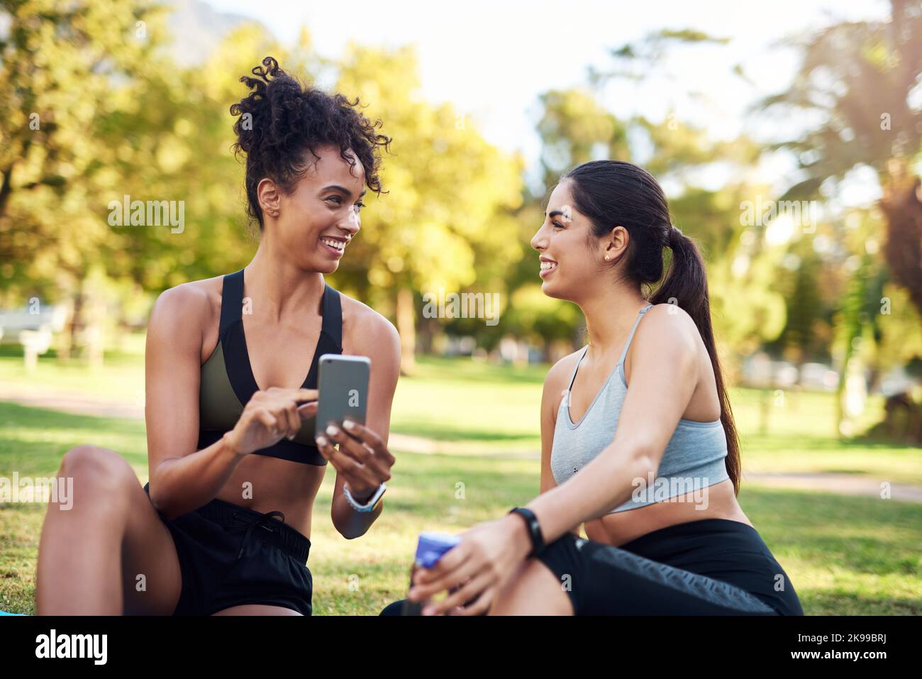 I think we deserve to take selfies. two attractive young women sitting next to each other while using a cellphone in the park. Stock Photo