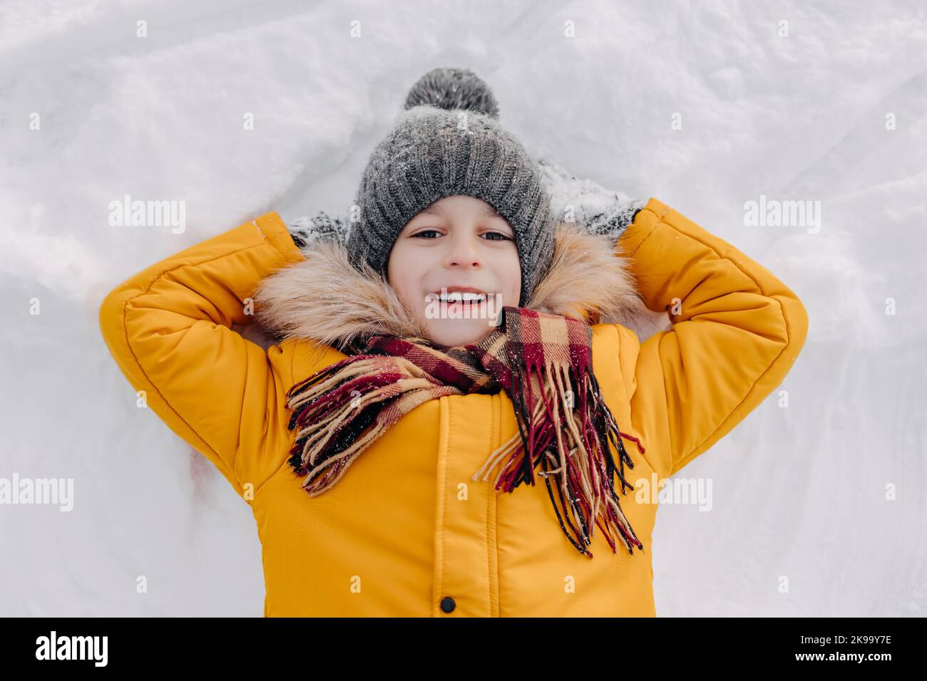 Portrait of happy eight years old kid lying on the snow. Smiling boy in warm clothing having fun outdoors. Concept of christmas holidays, active leisu Stock Photo