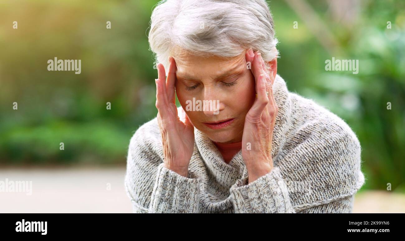 These headaches are irritating me. a stressed out elderly woman seated on a bench and holding her head in discomfort outside in a park. Stock Photo