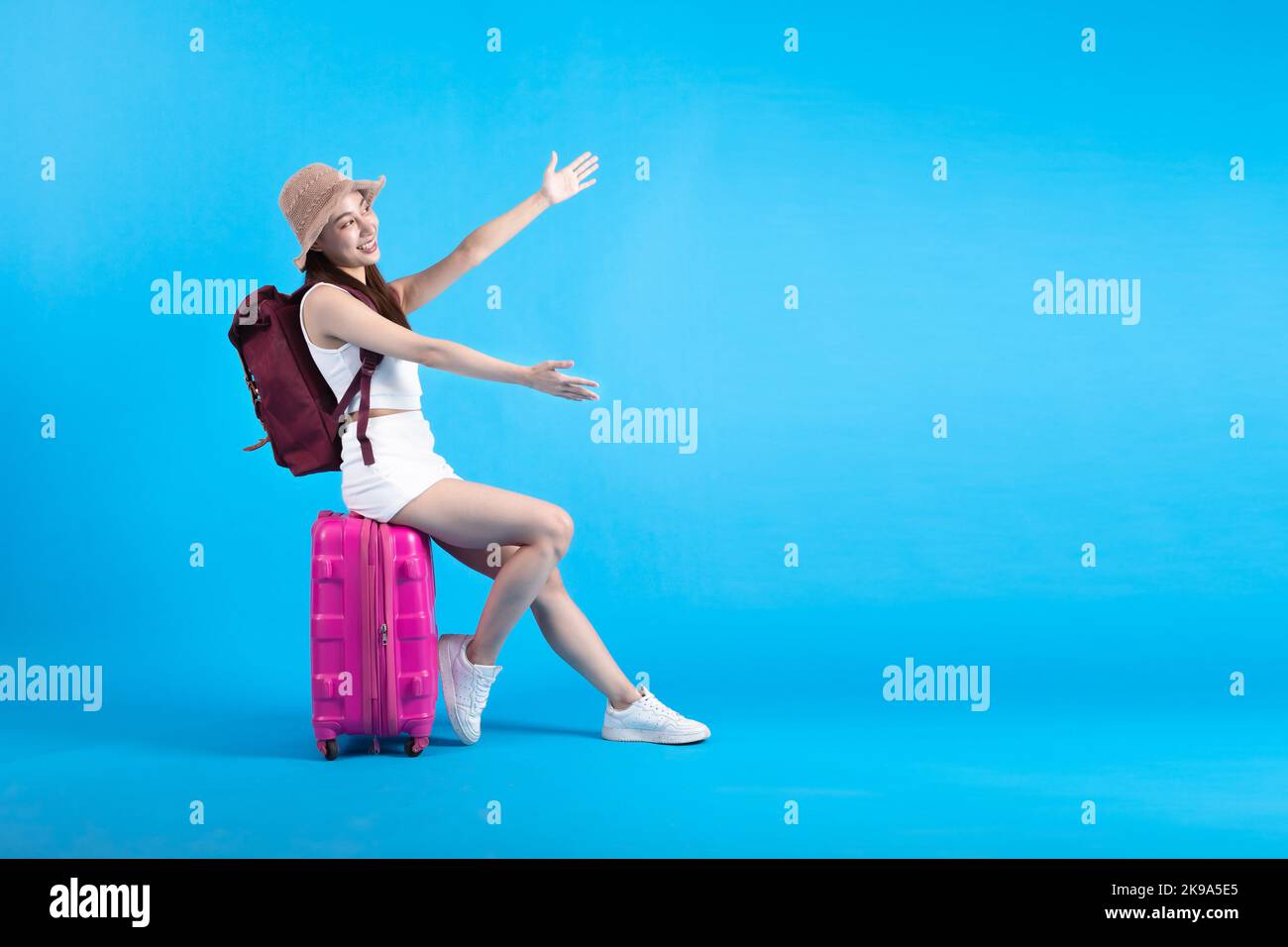 Happy Asian woman traveler sitting on suitcase luggage and showing the empty space isolated on blue background, Tourist Asia girl having exciting holiday trip, Full body length with copy space Stock Photo