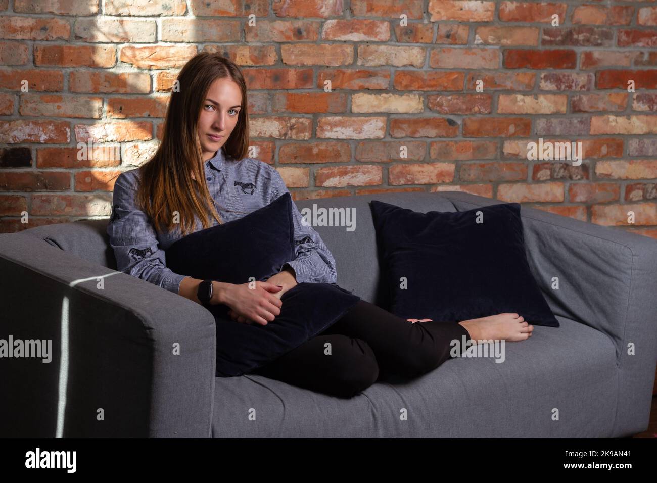 A woman in a blue shirt is on the couch, holding a pillow and looking into the camera Stock Photo