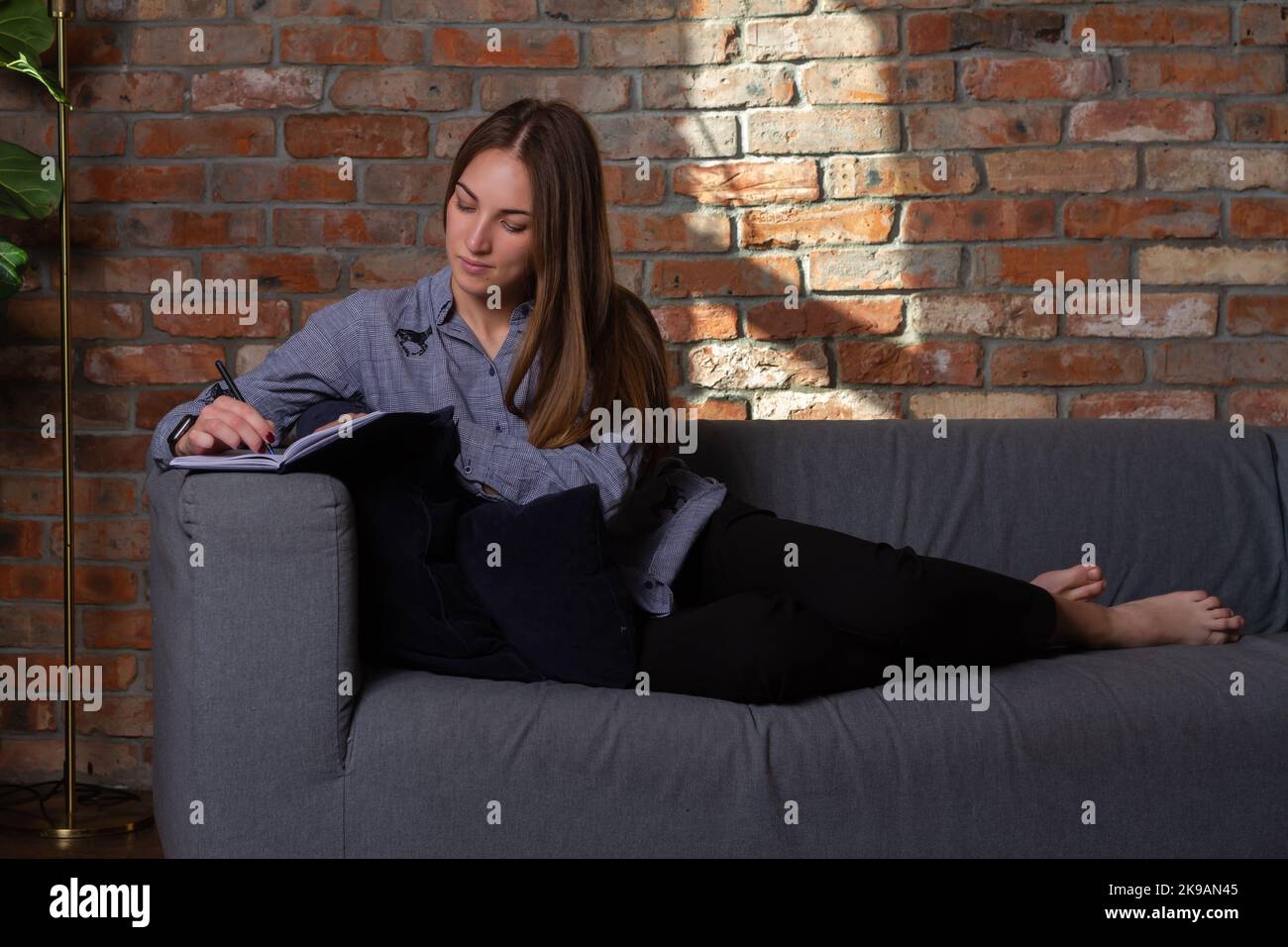 Woman in a blue shirt lying on the couch and writing in a notebook with a pen Stock Photo