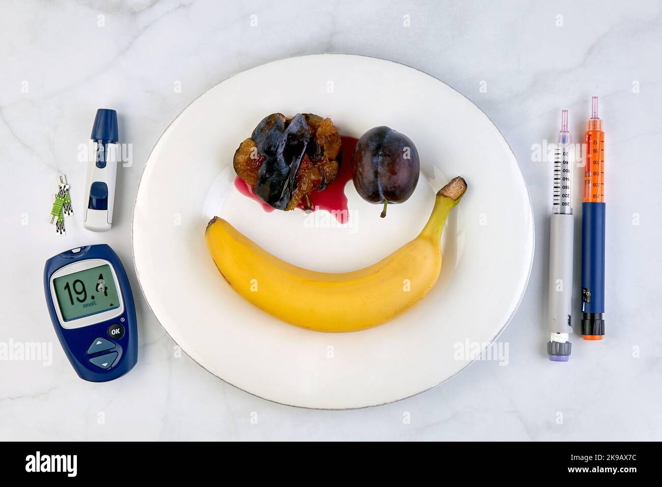 Banana and crushed plums in the form of a smiley face on a white plate, insulin pens and a glucometer with a very high blood glucose level Stock Photo