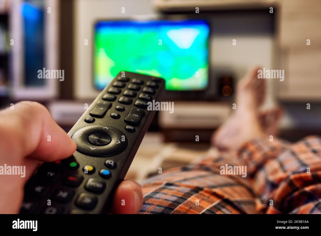 Person lying on bed with crossed legs and switching television channels on TV with finger on remote controller Stock Photo