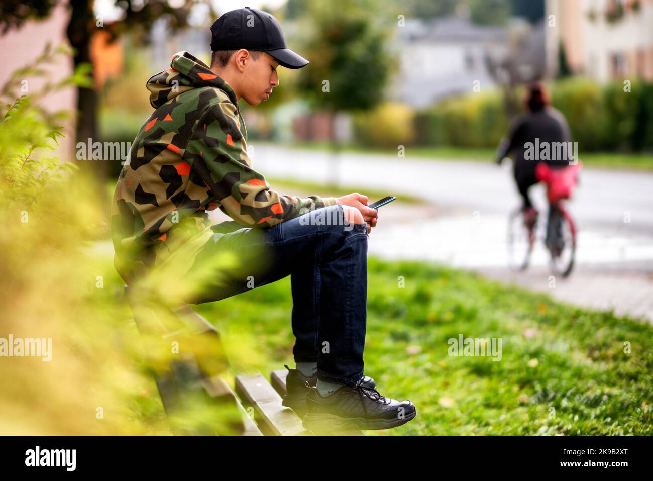 Young boy with black cap sitting on bench in city and using smarthphone. Stock Photo