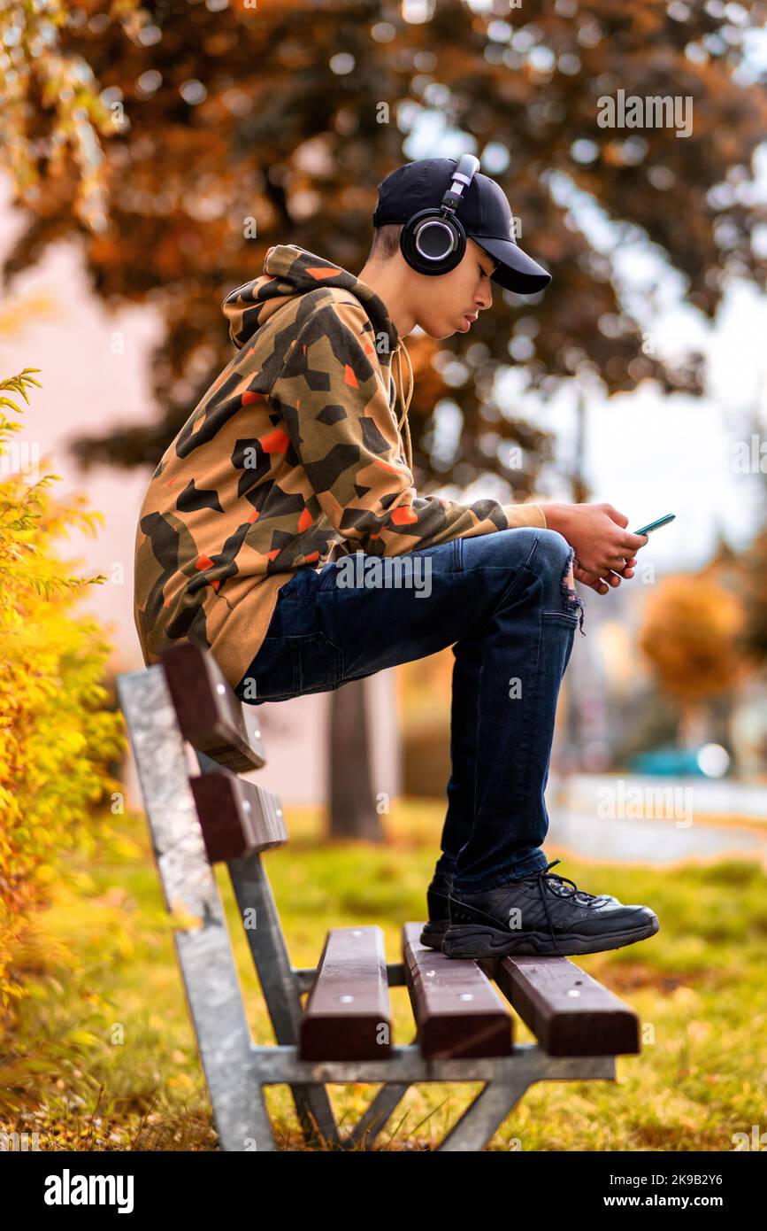Teenager boy with wireless headphones on head  sitting on bench in city and listening music Stock Photo