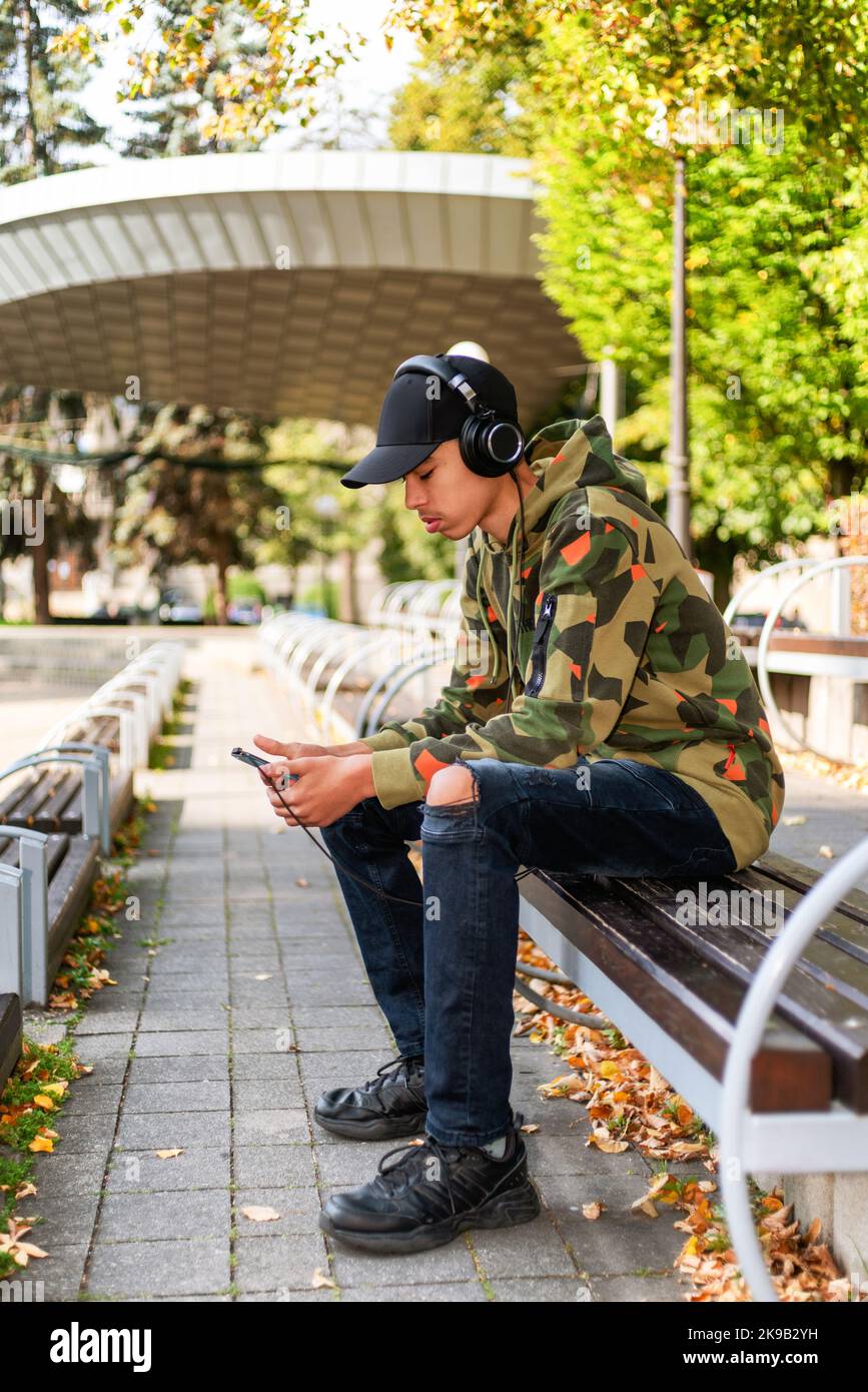 Young boy with black cap sitting on bench in park and using smarthphone. Stock Photo