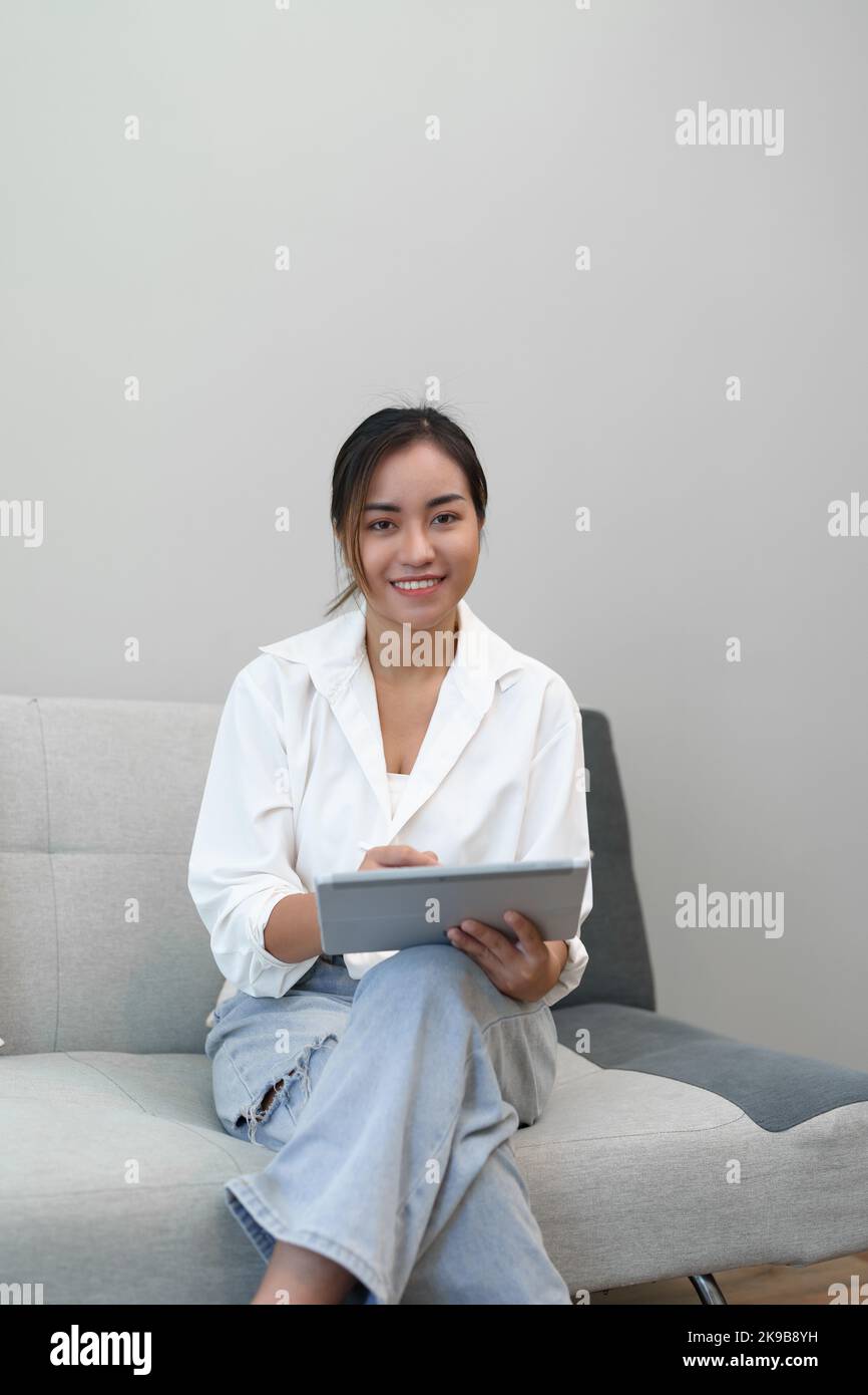 Portrait of a young Asian woman sitting on the sofa using a tablet ...
