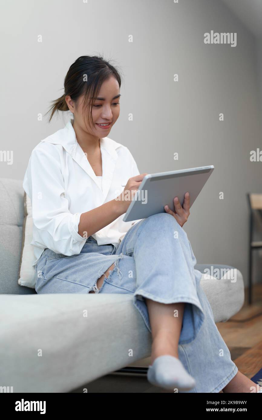 Portrait of a young Asian woman sitting on the sofa using a tablet ...