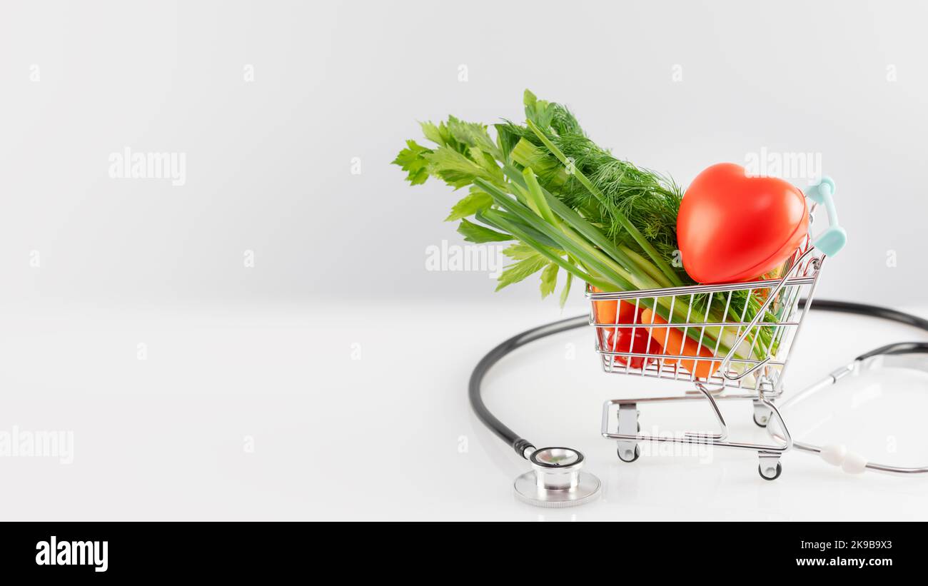 Health care banner with organic vegetables in a shopping cart with decorative heart and stethoscope on a white table with copy space. Concept of healt Stock Photo