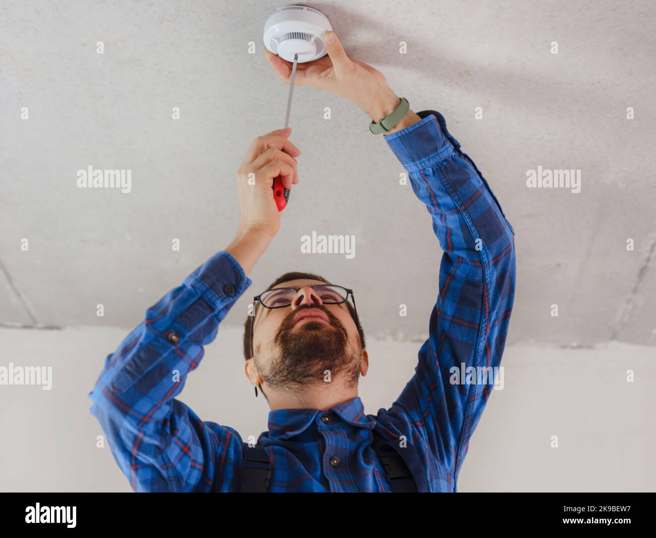 young electrician man in blue work suit installing smoke detector on the ceiling Stock Photo
