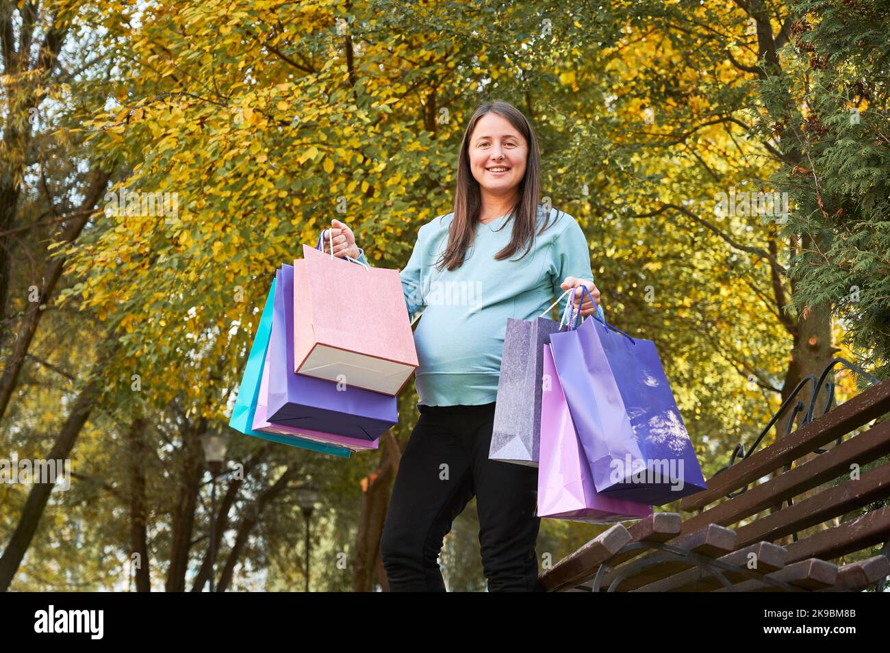 Half length portrait of cheerful pregnant woman smiling at camera after daytime shopping during awaiting BABY. Happy mother with paper bags enjoying leisure for preparing to appearance of toddler Stock Photo