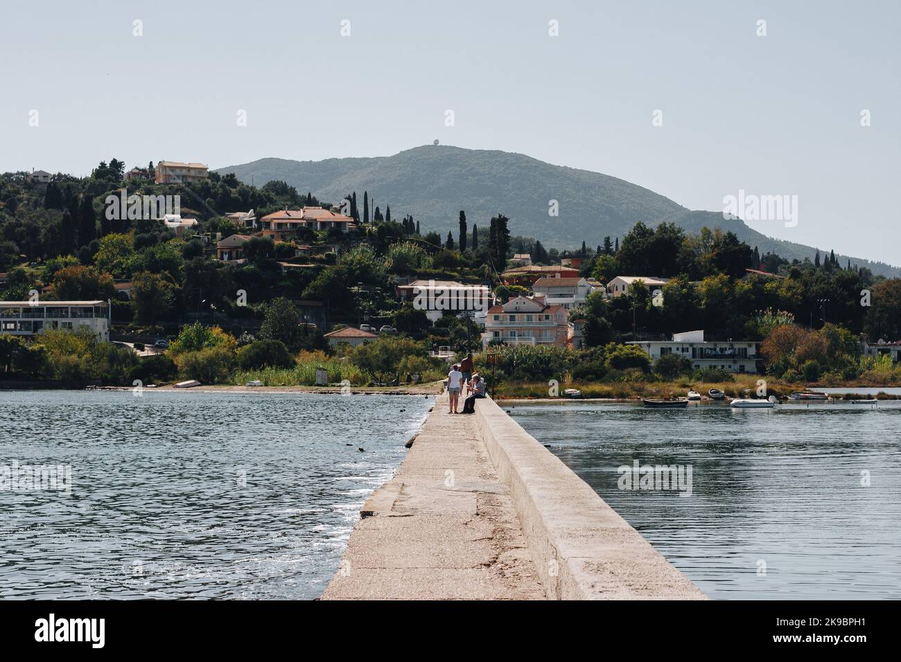 Corfu, Greece 2022.10.04. A hill with residential houses by the water with a pedestrian pier on which tourists sit Stock Photo