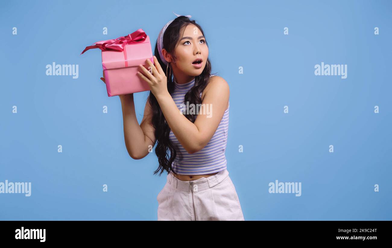 puzzled asian woman shaking wrapped gift box isolated on blue Stock Photo