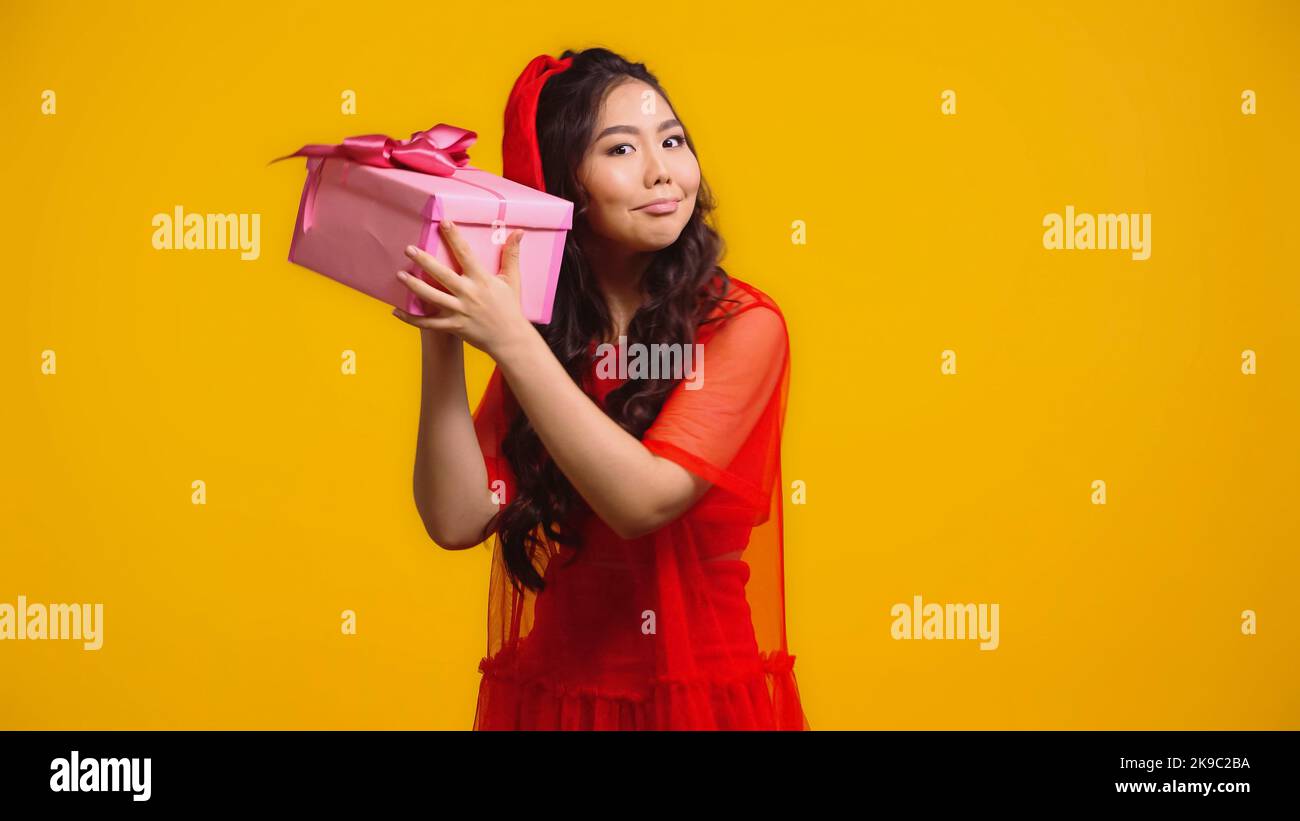 uncertain young asian woman shaking wrapped present isolated on yellow Stock Photo