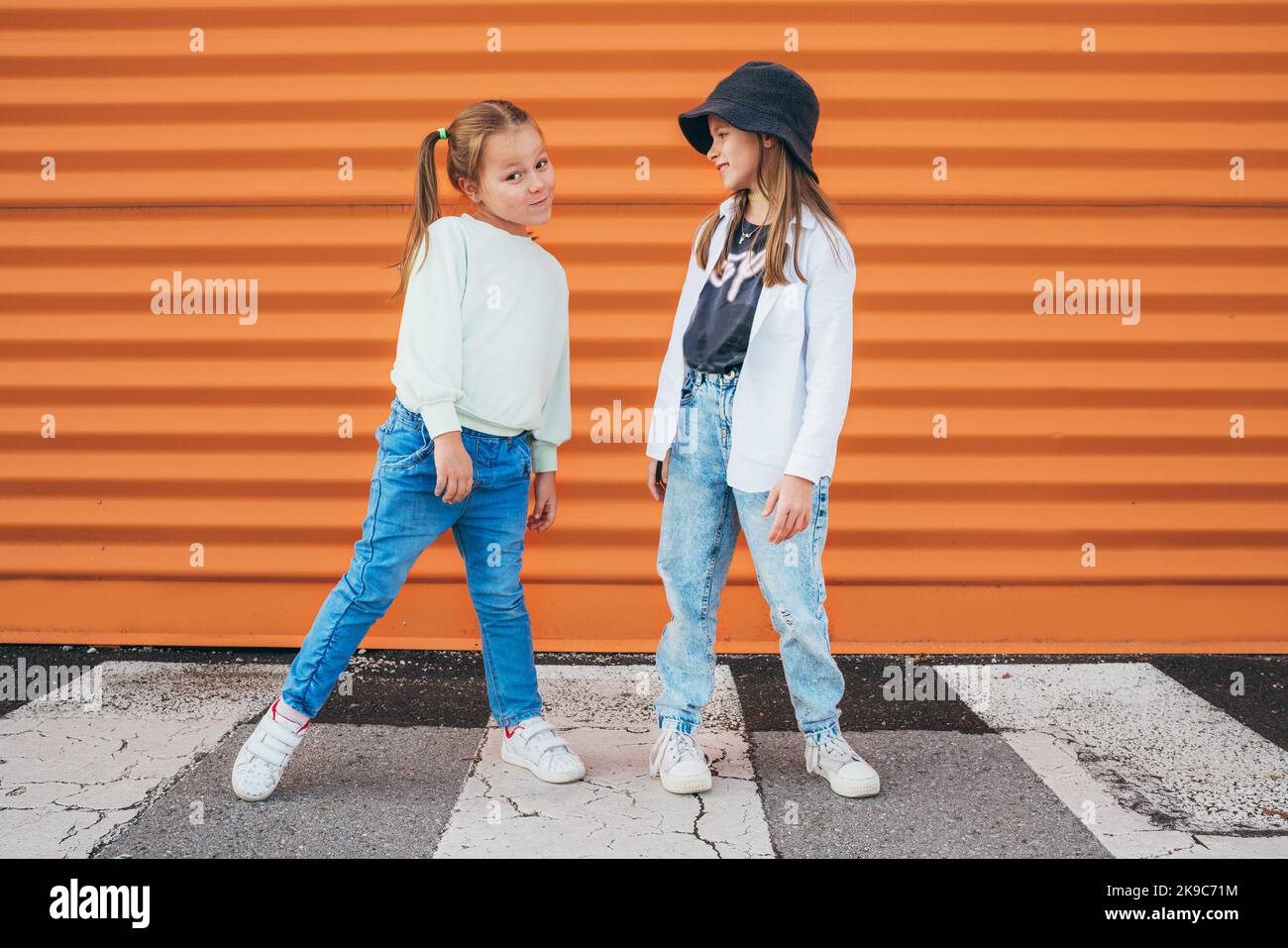 https://c8.alamy.com/comp/2K9C71M/two-little-girls-sisters-dressed-kids-fashion-vogue-style-clothes-posing-at-camera-on-the-city-pedestrian-asphalt-road-near-the-orange-wall-background-2K9C71M.jpg