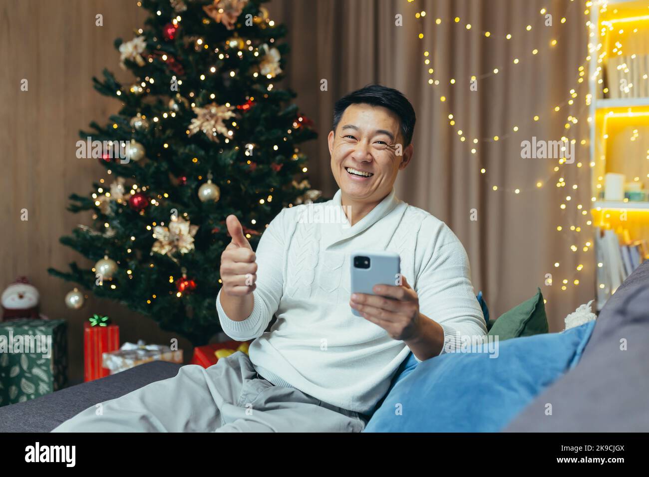 Asian man on christmas at home looking at camera and smiling, man celebrating new year and holidays, sitting on sofa in living room holding phone. Stock Photo