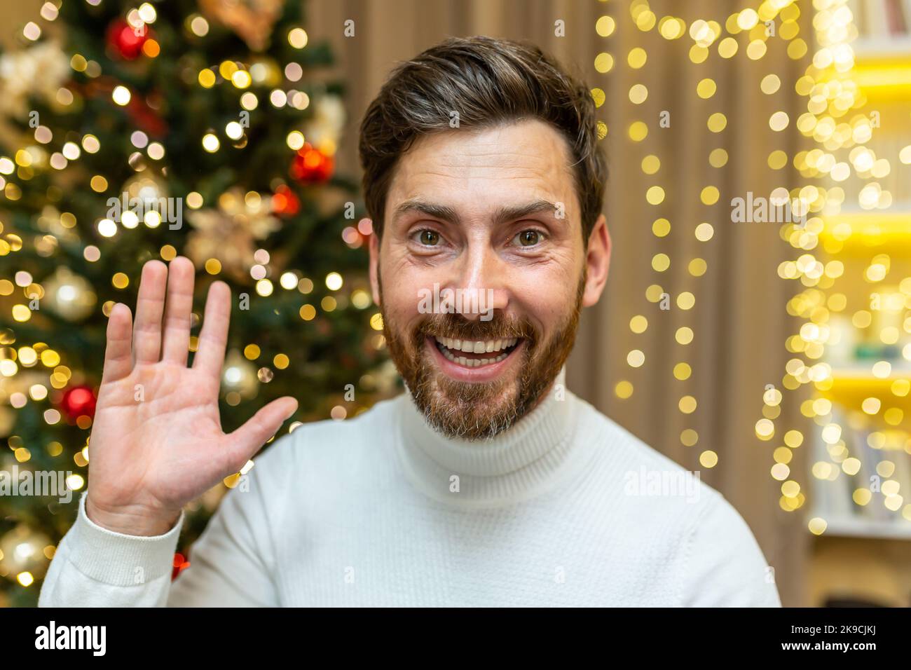 Online greetings video call, Christmas man looks into web camera and smiles and waves while sitting on sofa at home celebrating New Year holidays. Stock Photo