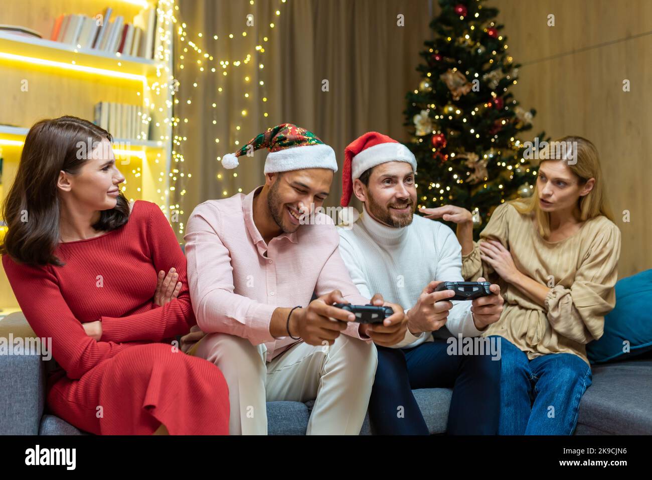 New Year's party group of four diverse friends having fun relaxing and celebrating on Christmas holidays, guests sitting on sofa men and women playing video games on joystick consoles. Stock Photo