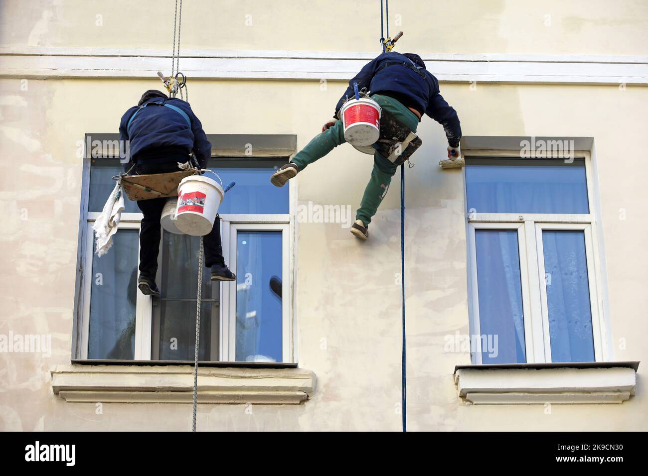 Workers paints the building wall. Painters hanging on a cables with paint buckets, steeplejack repairing house facade with roller brush Stock Photo