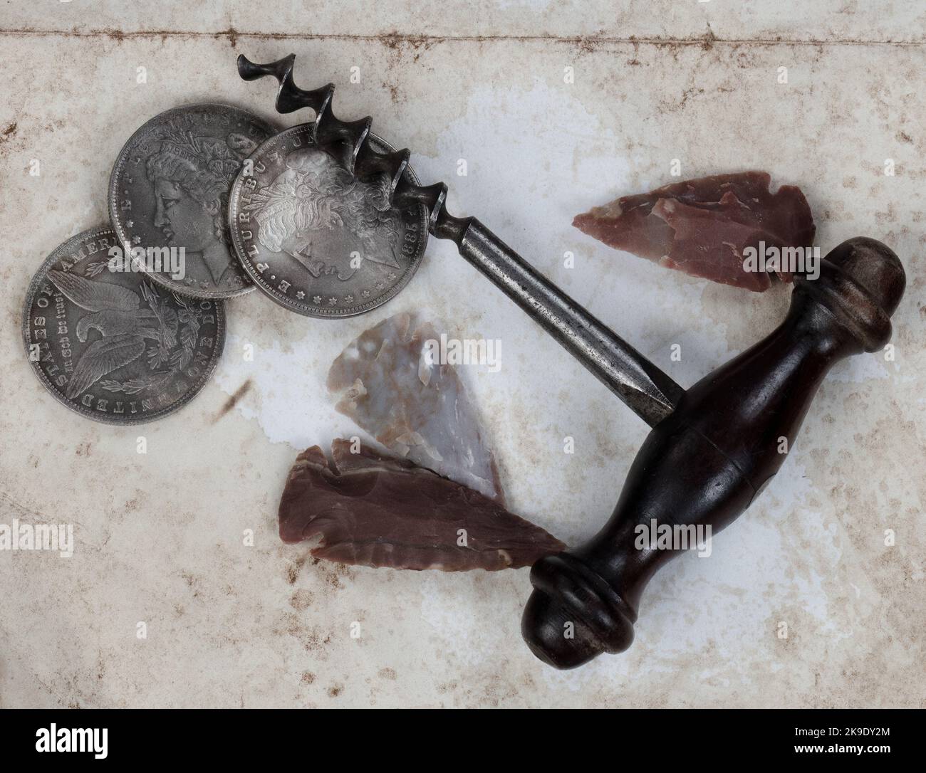 Unique vintage collection of antique corkscrew, native American arrowheads, and silver dollar coins on fade paper background in close up view Stock Photo