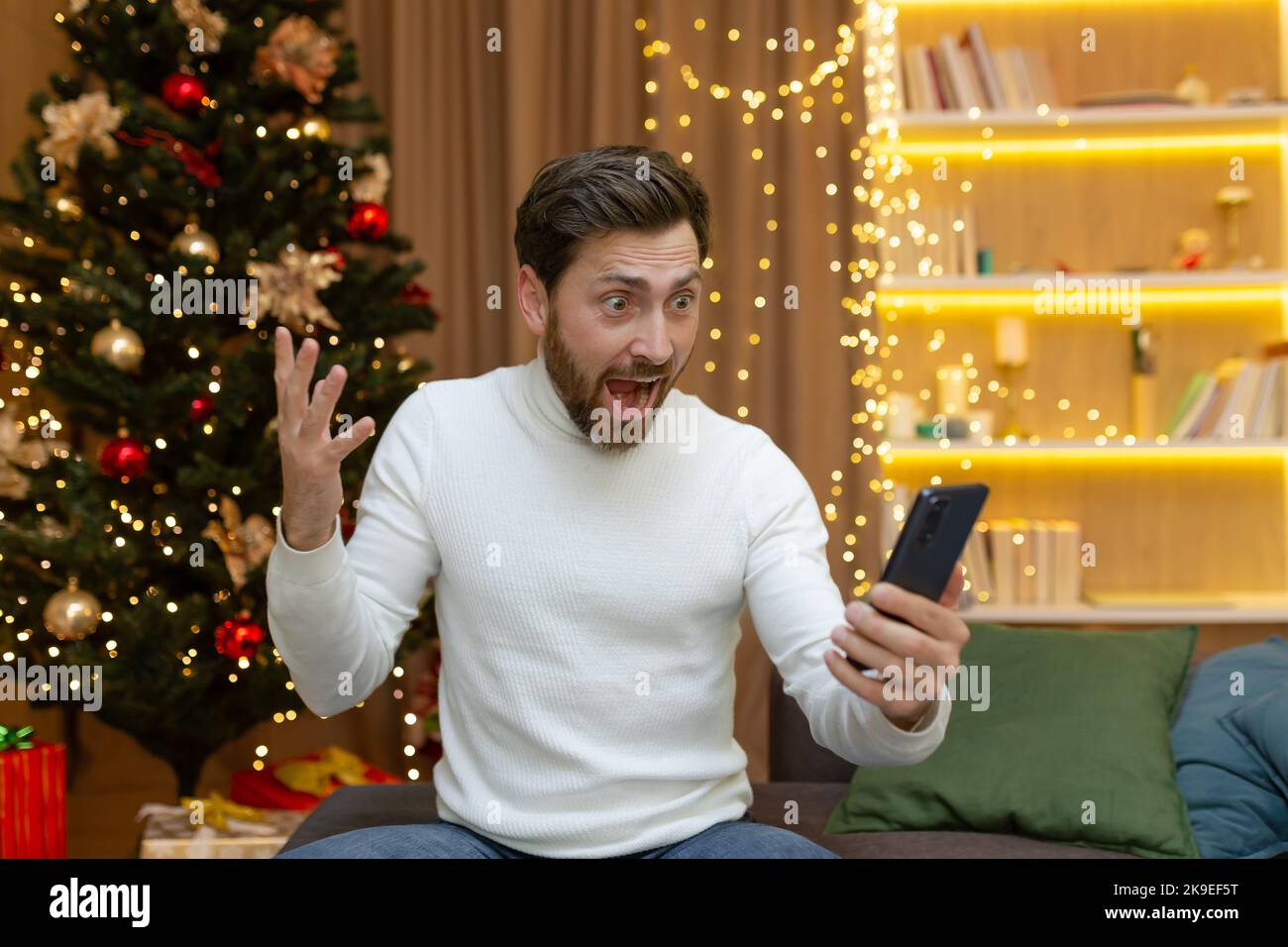 A happy young handsome man is sitting on a sofa in a New Year's decorated apartment. He holds the phone in his hands, rejoices, finds good pre-Christmas and New Year's discounts on gifts Stock Photo