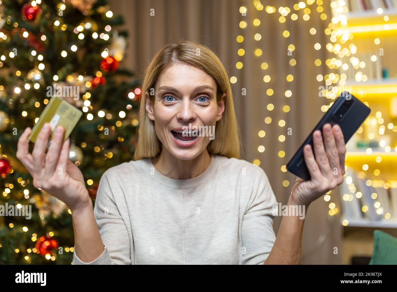 Portrait of happy online shopper woman, christmas woman looking at camera and smiling happy holding bank credit card and phone in hands, sitting on sofa near tree celebrating new year. Stock Photo