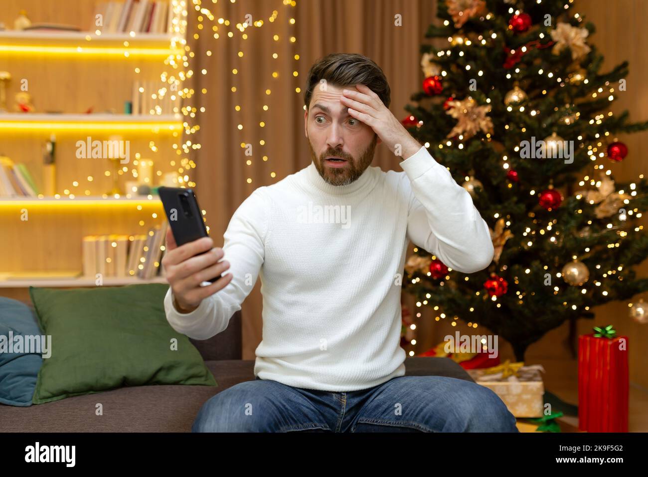 Upset and disappointed christmas man reading online messages and notifications from smartphone, sitting at home on christmas sofa during new year holidays. Stock Photo