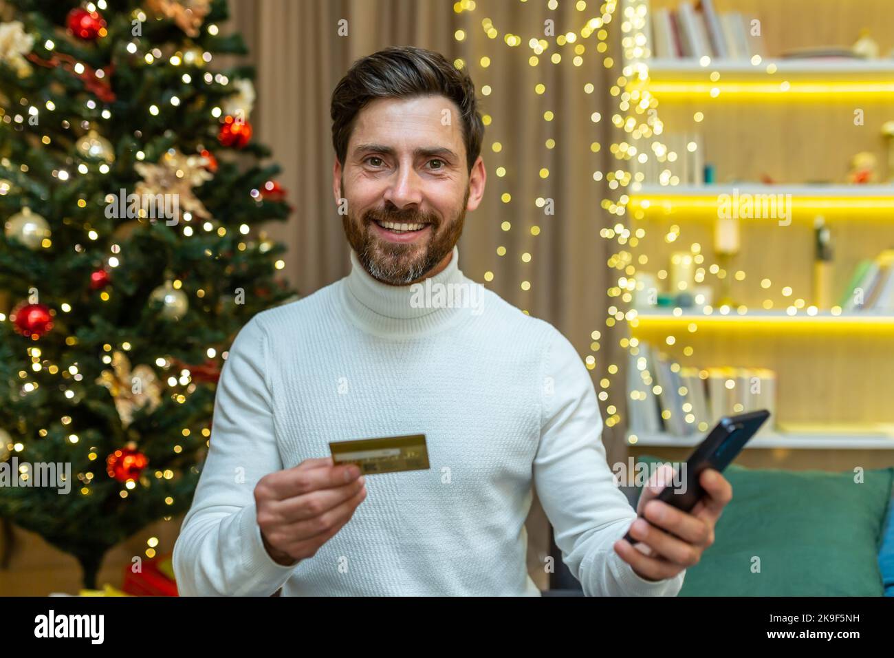 Christmas man looking at camera and smiling, holding smartphone and bank credit card for online shopping in online store, sitting on sofa in living room on New Year holiday. Stock Photo