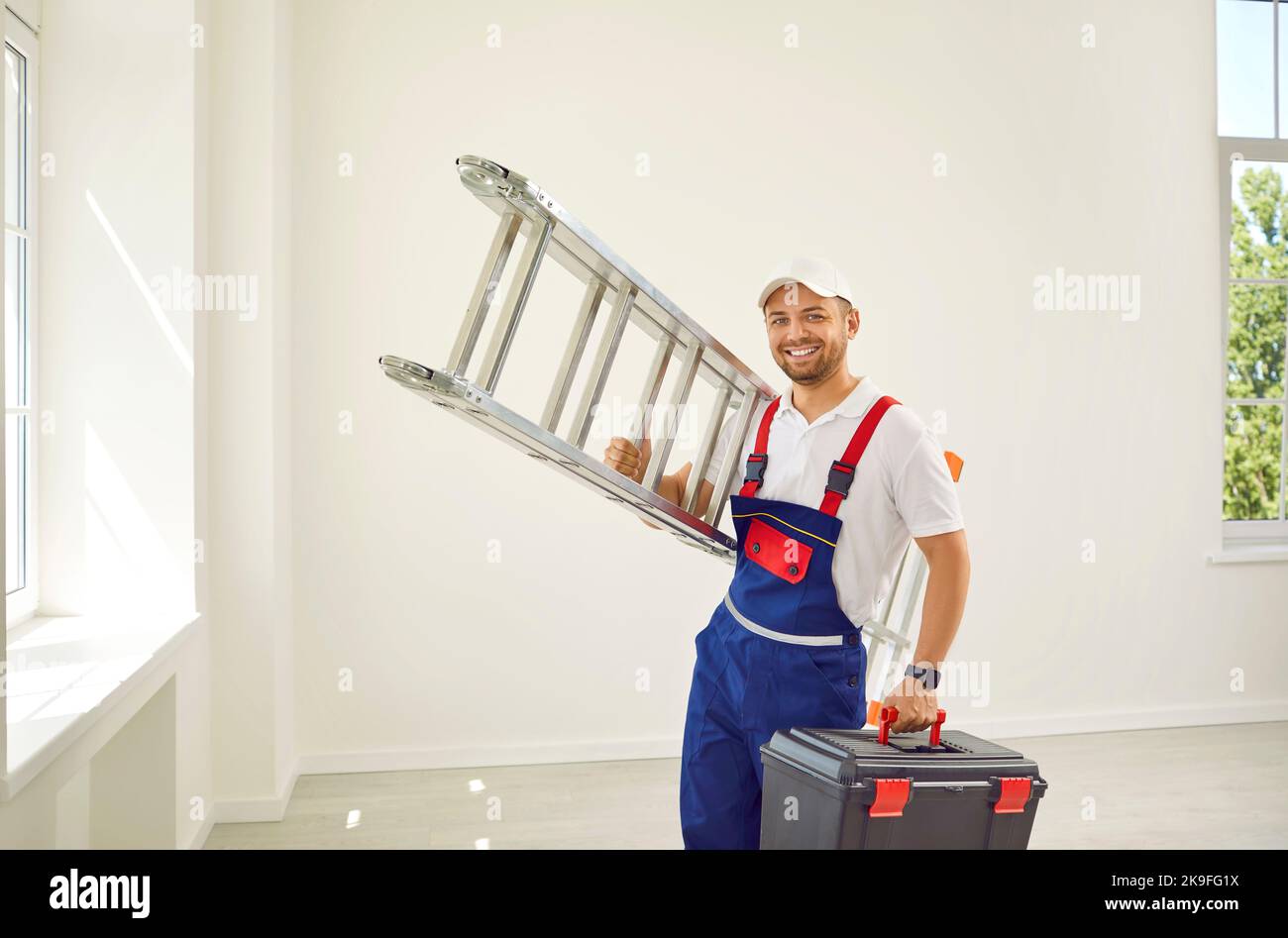 Happy repairman standing in the room, holding a ladder and a toolbox and smiling Stock Photo