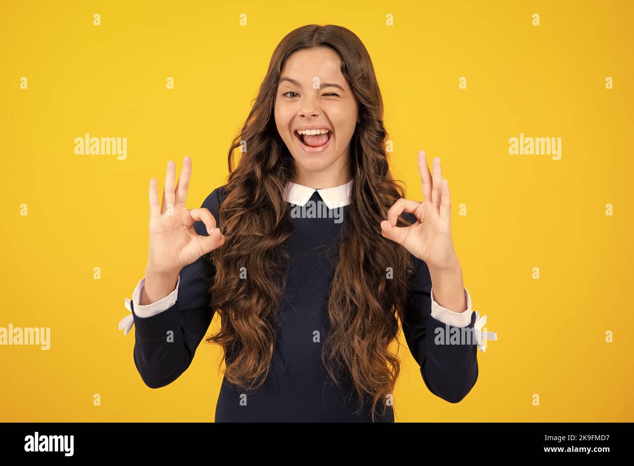 Excited face. Smiling teen girl, young teenager showing ok hand sign and winking looking at camera isolated on yellow studio background. Amazed Stock Photo