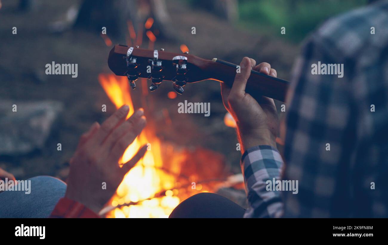Close-up shot of male hands playing the guitar and female hands moving in dance with burning campfire in background. Camping, music and adventurous people concept. Stock Photo