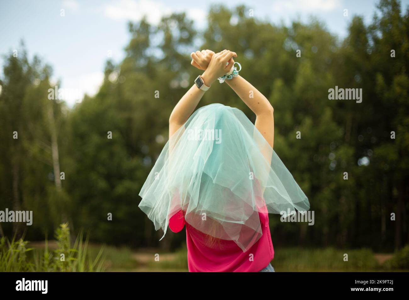 Blue veil on head. Bridesmaid's clothes. Lightweight fabric in summer. Girl in park. Hands raised up. Pink t-shirt. Stock Photo