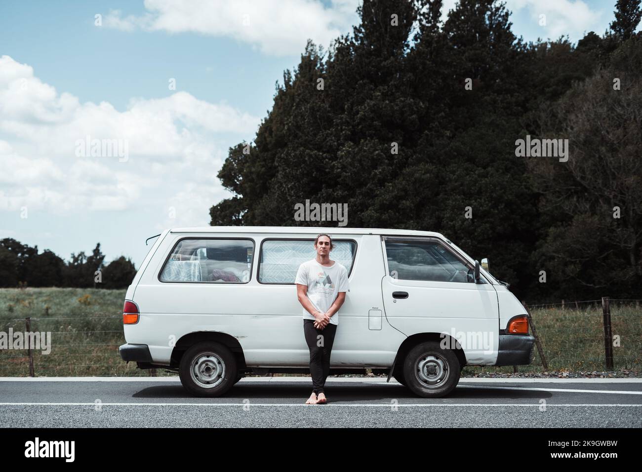 barefoot caucasian young man dressed in a t-shirt and black pants standing cross-legged leaning against a white van parked on the road, rotorua, new Stock Photo