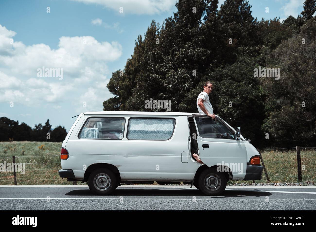 barefoot disheveled caucasian boy standing perched on the right edge of a white van leaning against the open door looking at camera, rotorua, new Stock Photo