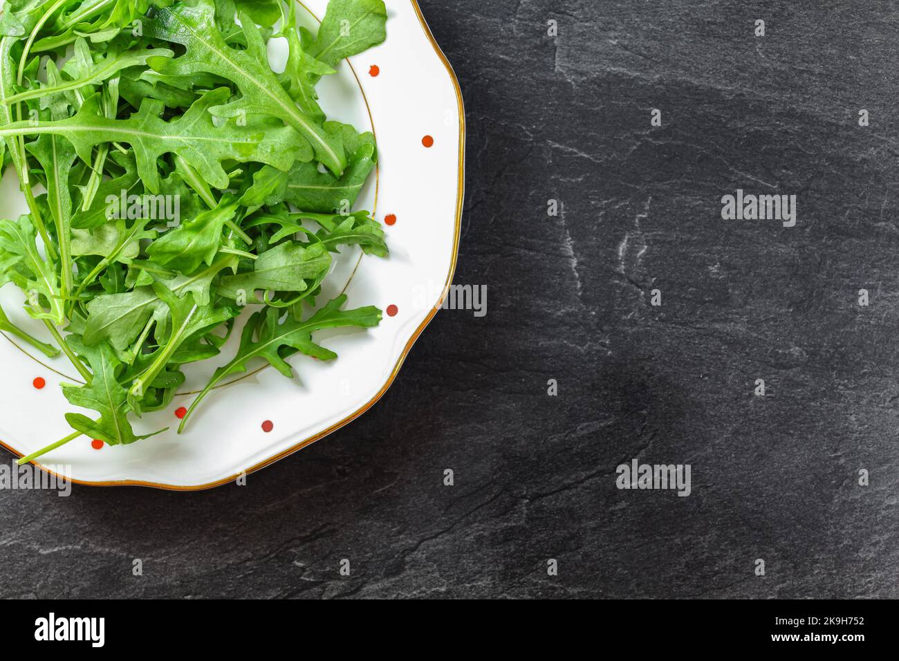 Fresh green arugula or garden rocket leaves on white plate with red dots, black table space for text right side under. Healthy greens and salads Stock Photo