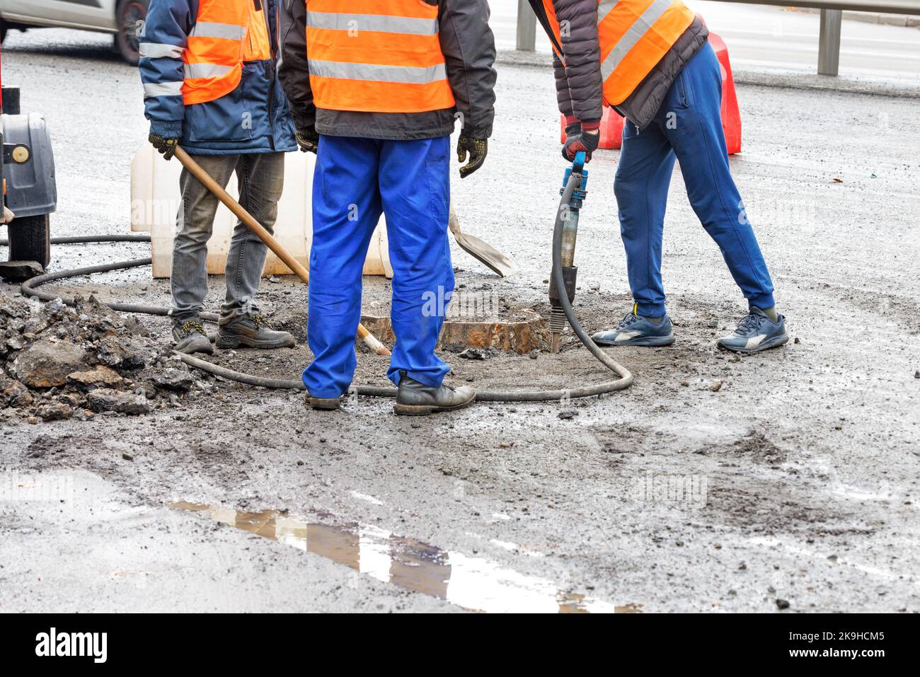 A team of road workers remove old asphalt around a sewer manhole with a jackhammer and shovel for later repair. Copy space. Stock Photo