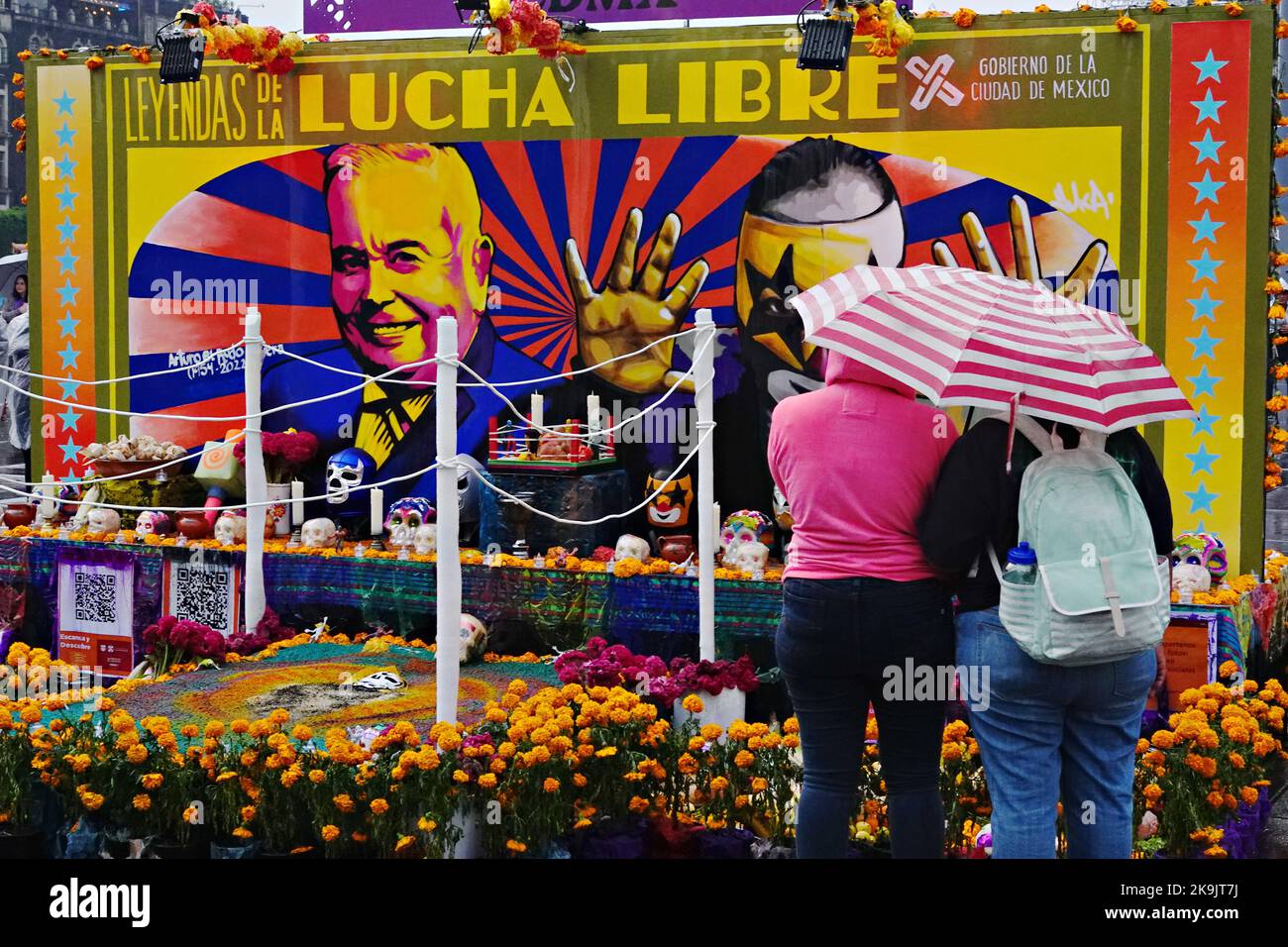 Mexico City, Mexico. 28th Oct, 2022. Two people hide under and umbrella during an afternoon thunderstorm as they view a Ofrenda or altar honoring Mexican wrestling during the Megaofrenda of Zocalo festival to celebrate the start of the Day of the Dead holiday at the Plaza de la Constitucion, October 28, 2022 in Mexico City, Mexico. Credit: Richard Ellis/Richard Ellis/Alamy Live News Stock Photo