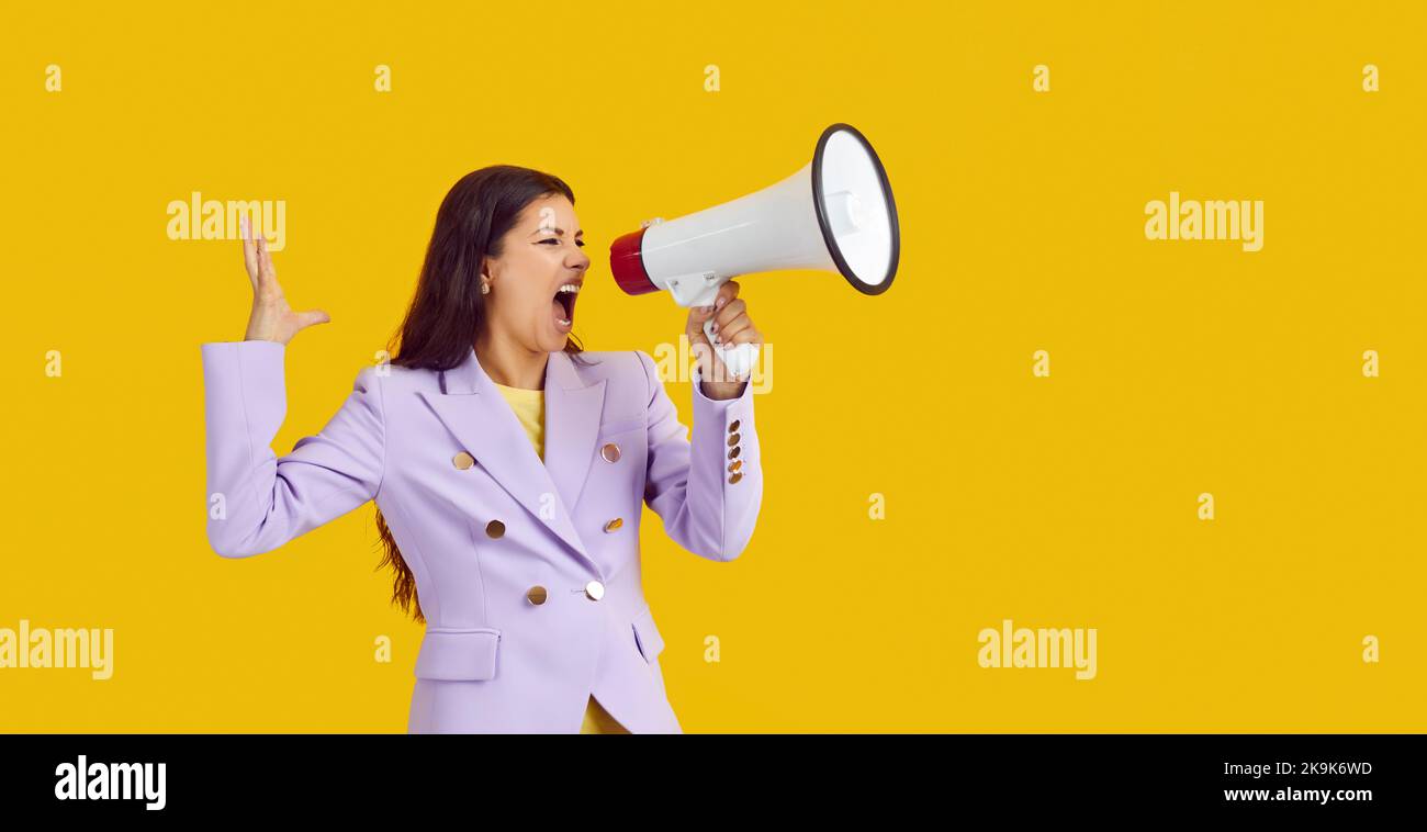 Emotional female activist with megaphone makes loud message on yellow background. Stock Photo