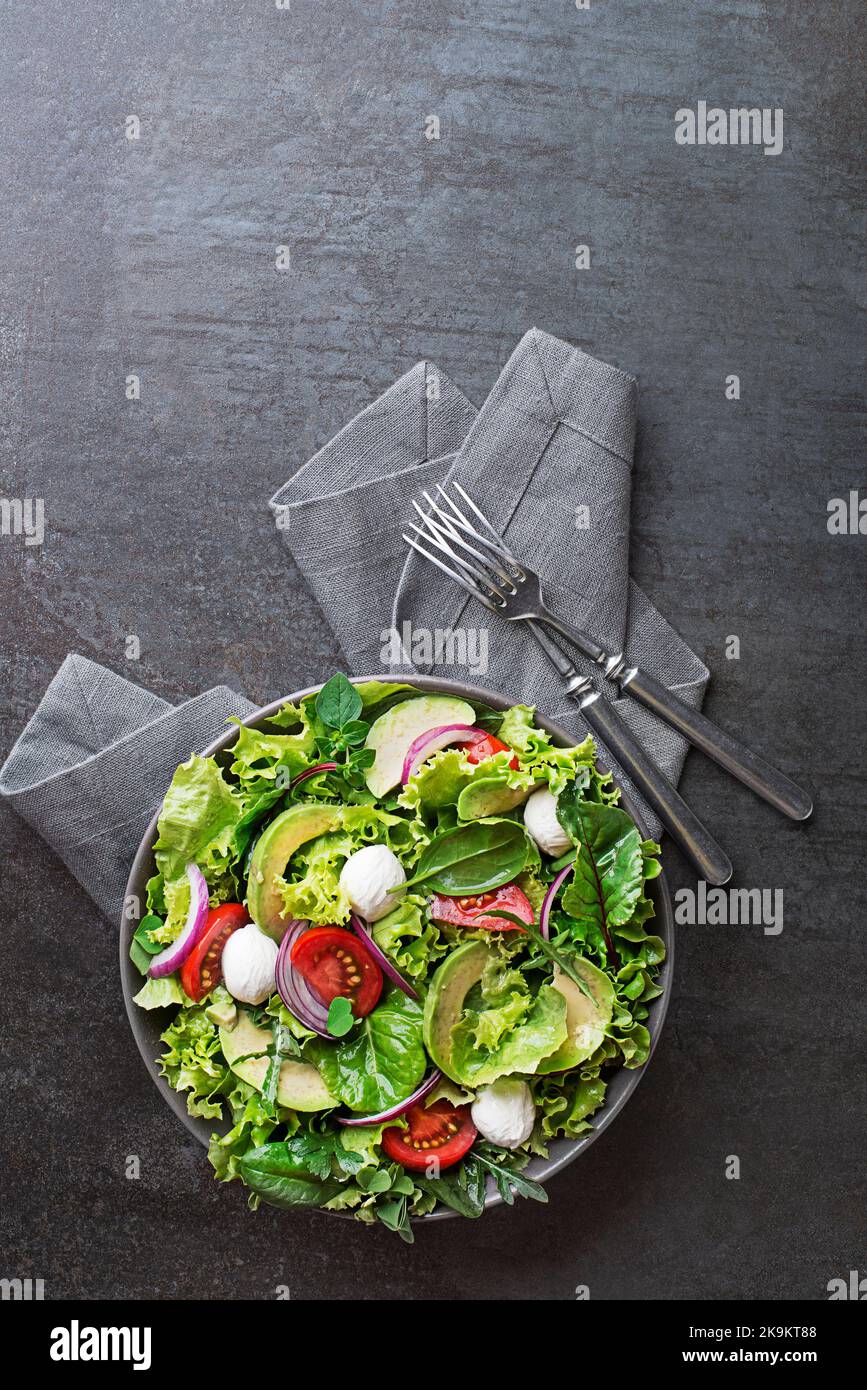 Healthy green salad with avocado, mozzarella cheese and fresh vegetables on dark background Stock Photo