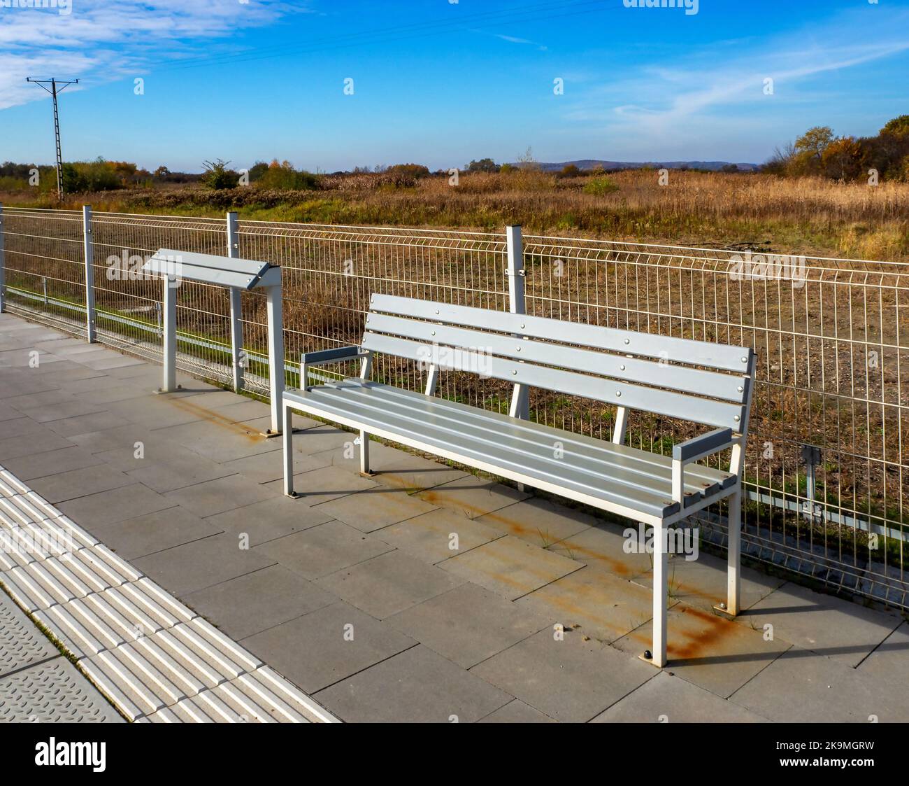 Traditional and leaning bench or lean bar (no established name yet) on the platform of a railway station. Leaning stand dries much faster after the ra Stock Photo
