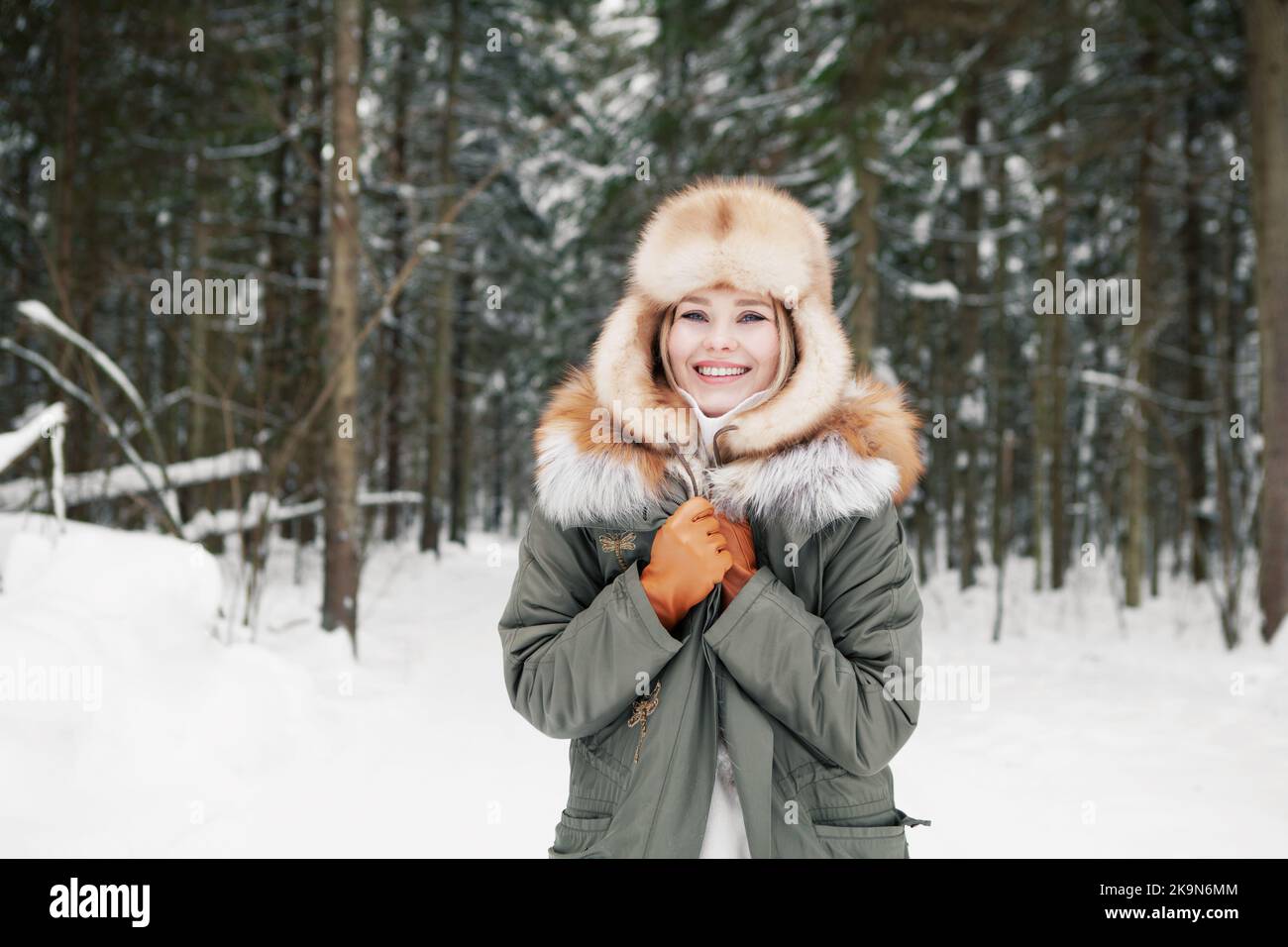 Smiling young woman in khaki parka, ear flaps ushanka hat and leather gloves at snowy coniferous forest background. Happy blonde model with beautiful Stock Photo