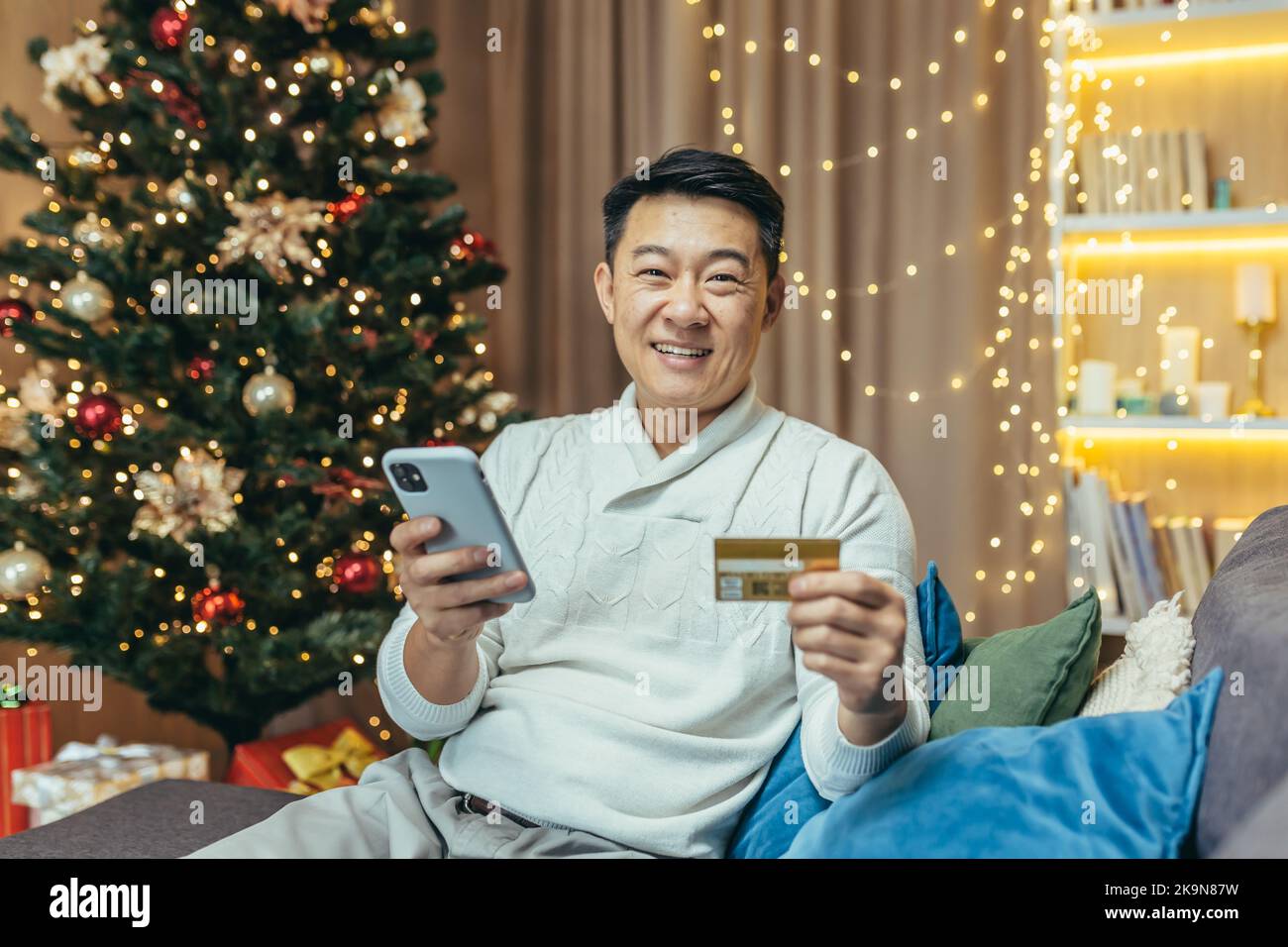 Happy Asian online shopping on new year Christmas eve buying and choosing gifts for friends in online store, portrait of happy man home looking at camera and smiling with bank credit card and phone Stock Photo