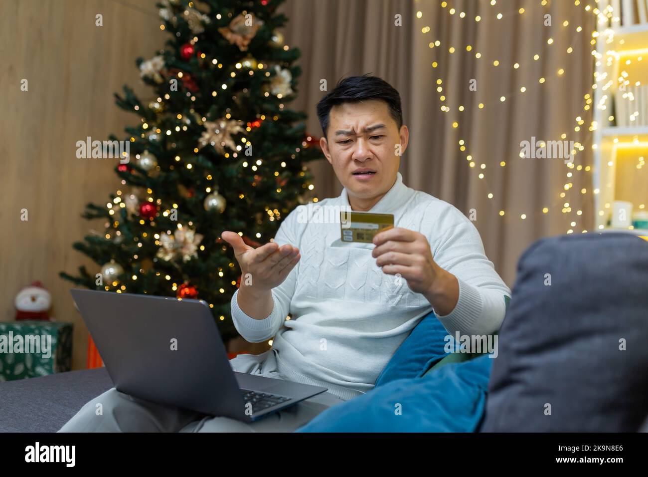 Upset christmas man with laptop trying to make online purchase in online store, asian man holding bank card received error and fraud message, sitting on couch for new year. Stock Photo