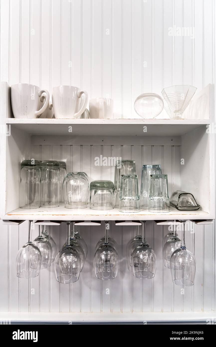 Closeup of modern kitchen room interior design white cabinet shelves with white cups, upside down hanging empty wine glasses rack for storage Stock Photo