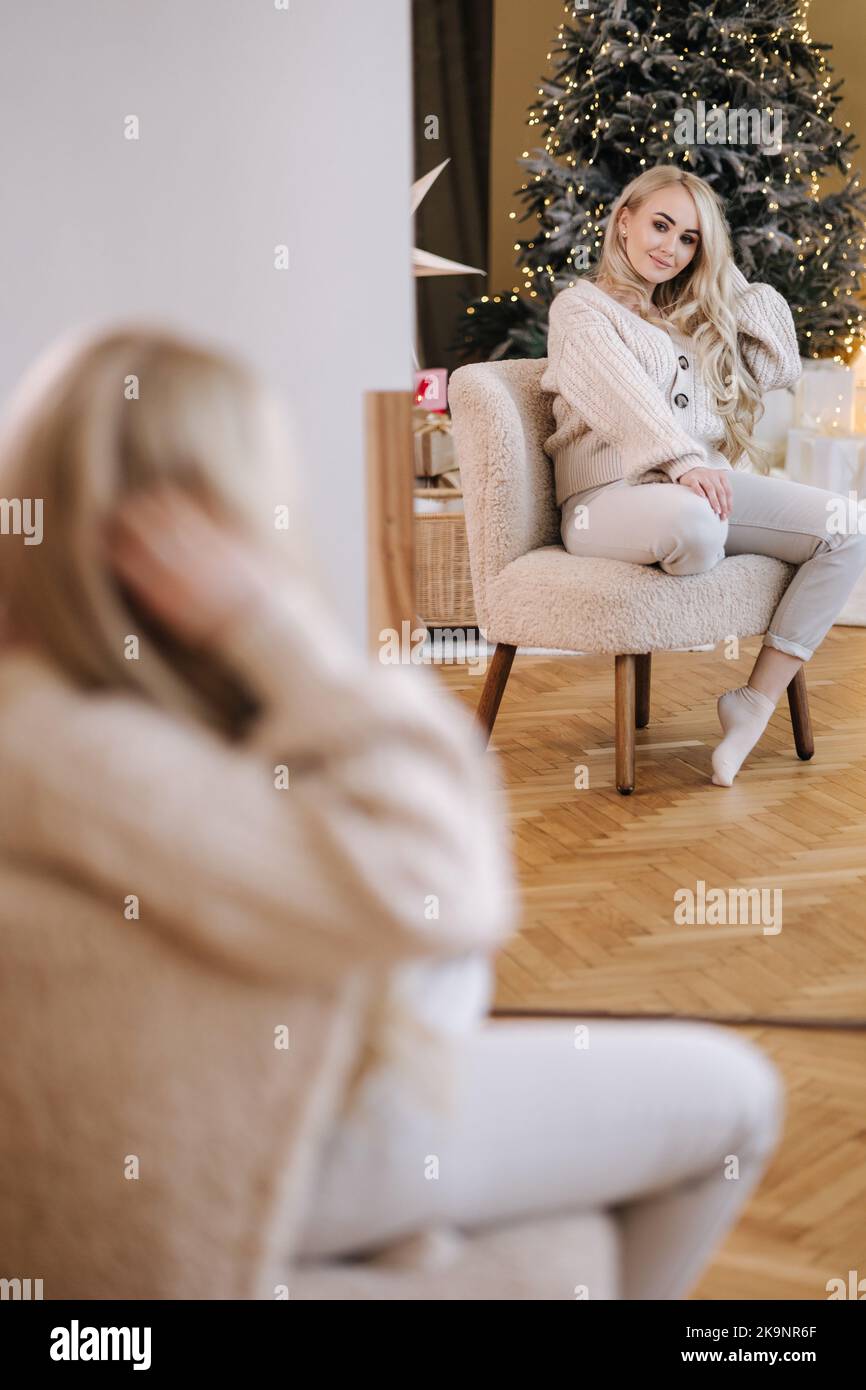 Attractive young blond hair woman in white knitted suit sitting on white chair in front of Christmas tree. Winter holidays Stock Photo