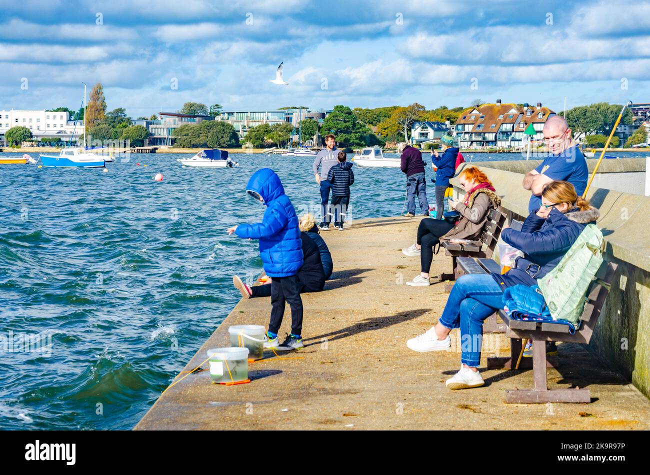 A family sit on benches and watch as a child enjoys crab fishing on the quay at Mudeford near Christchurch in Dorset, UK Stock Photo