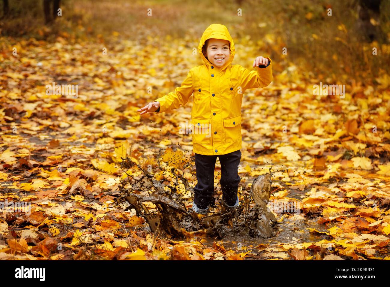 happy child boy in yellow waterproof raincoat and rubber boots jumping in puddle Stock Photo