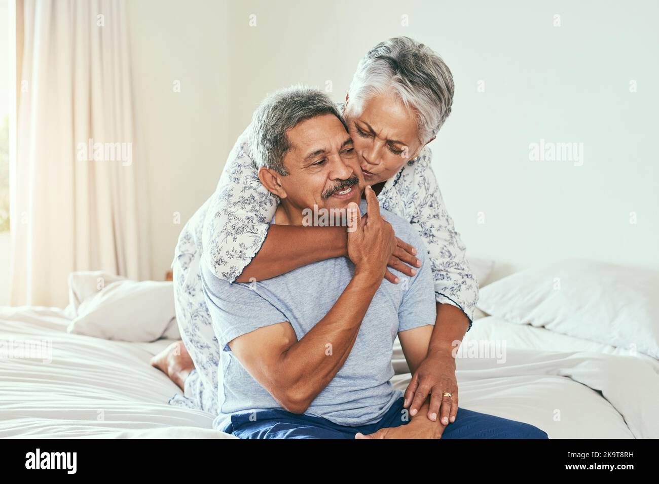 What a nice surprise. a cheerful mature man being held by his wife and receiving a kiss on the cheek while they both sit on the bed at home. Stock Photo